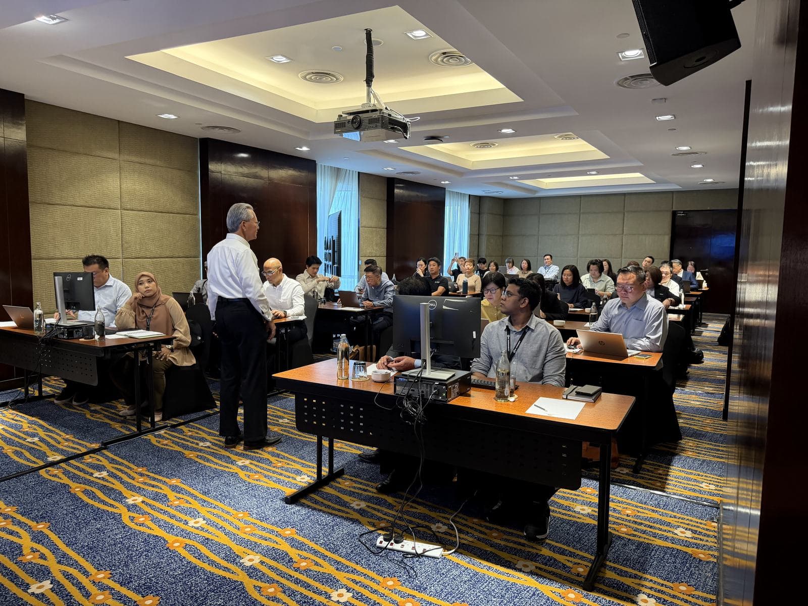 Presenter standing at the front of a hotel ballroom workshop with participants at desks and laptops