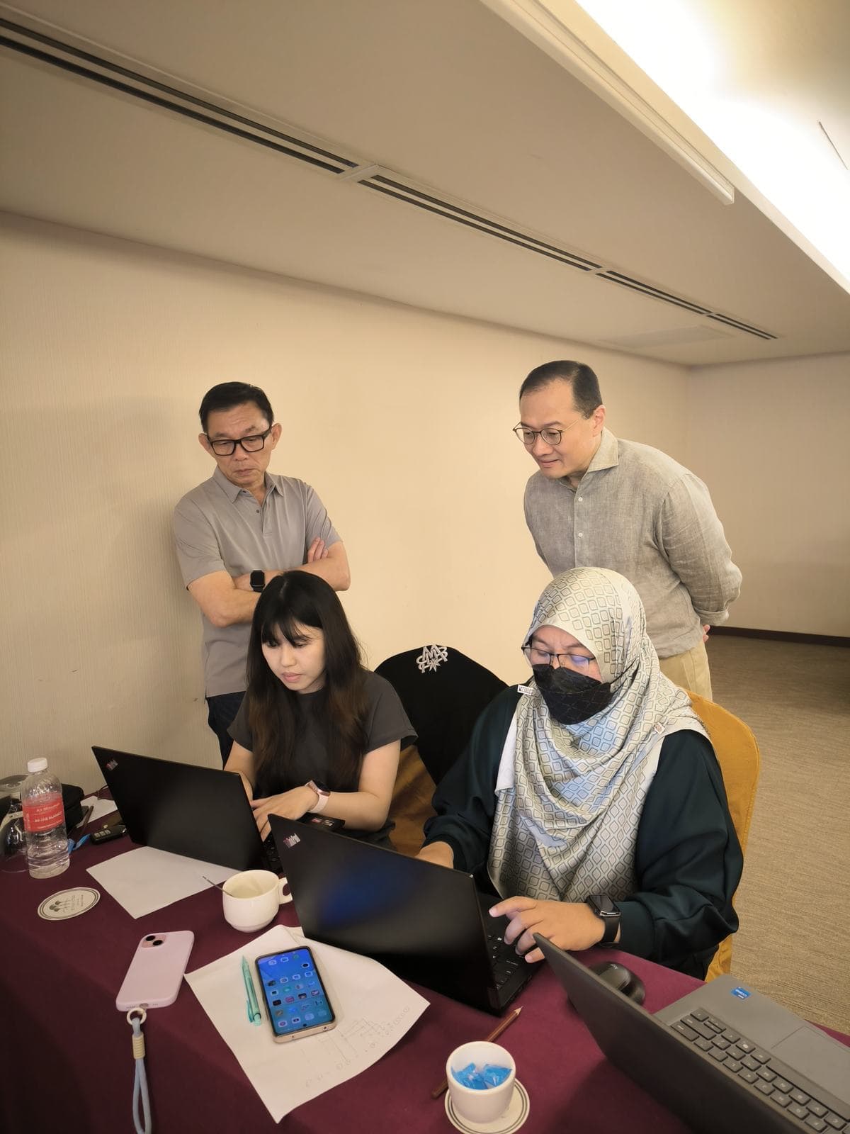 Facilitators coaching two participants at a laptop during a hands-on workshop in a hotel function room