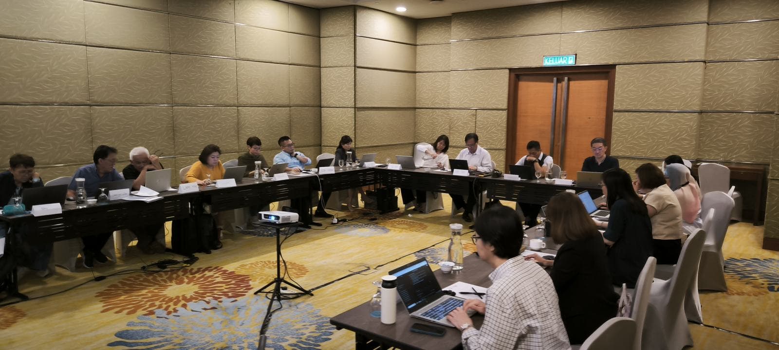 Senior participants seated around a U-shaped table with laptops and a projector in a hotel function room workshop