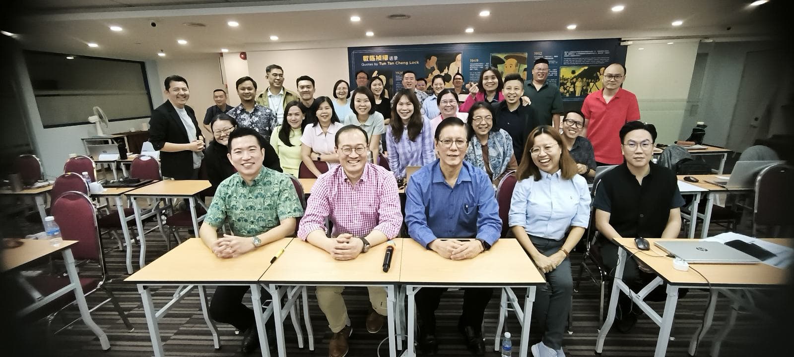 Group photo of a full classroom of workshop participants smiling for the camera, with three people seated at the front table