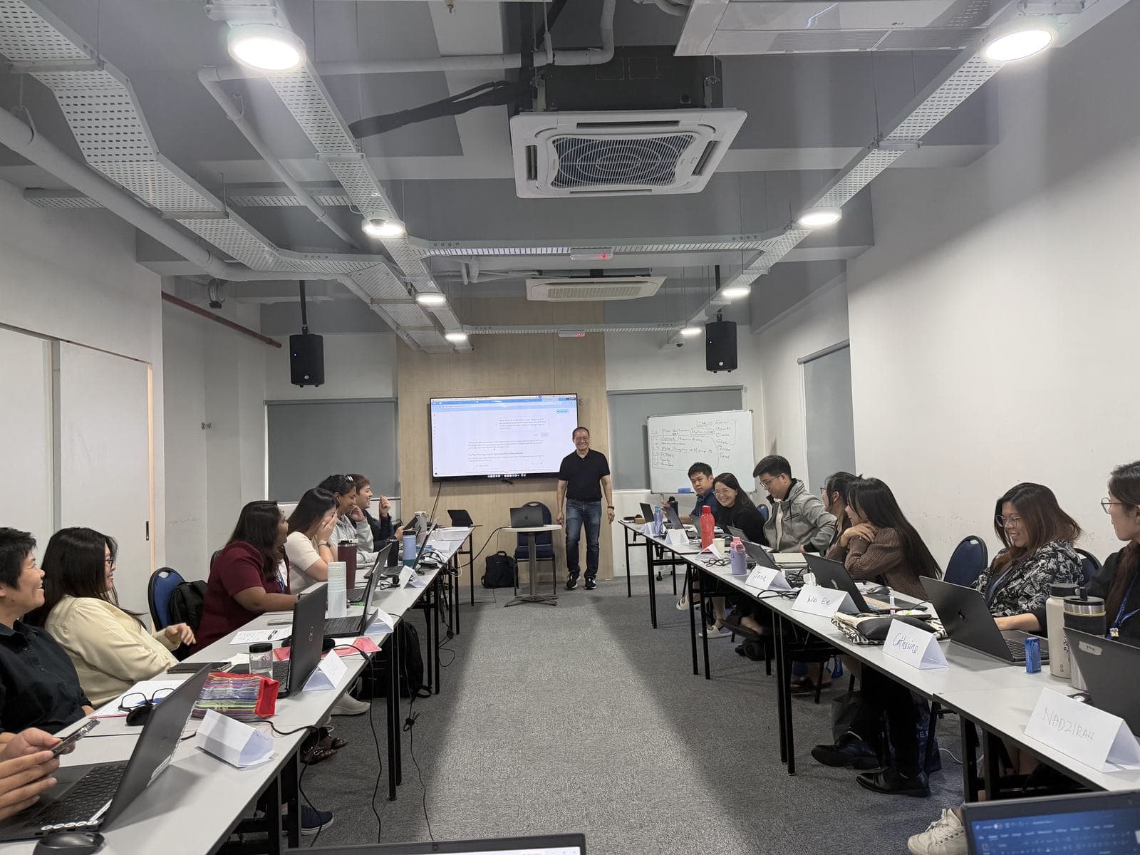 Facilitator presenting to participants seated at long tables with laptops in a training room
